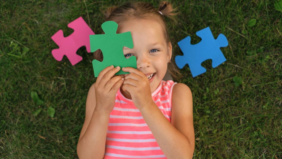 Preschool girl is lying on the grass on the lawn covering her face with puzzle pieces laughing merrily.