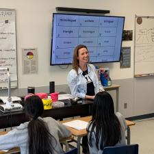 Female teacher stands at the front of the class as two students can be seen sitting at their desks looking at her