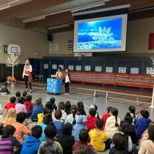 Assembly with students sitting on the ground, Principal standing at the front, with two students standing at a podium ready to speak.