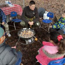 Students and one staff sitting around a gas firepit roasting marshmellows